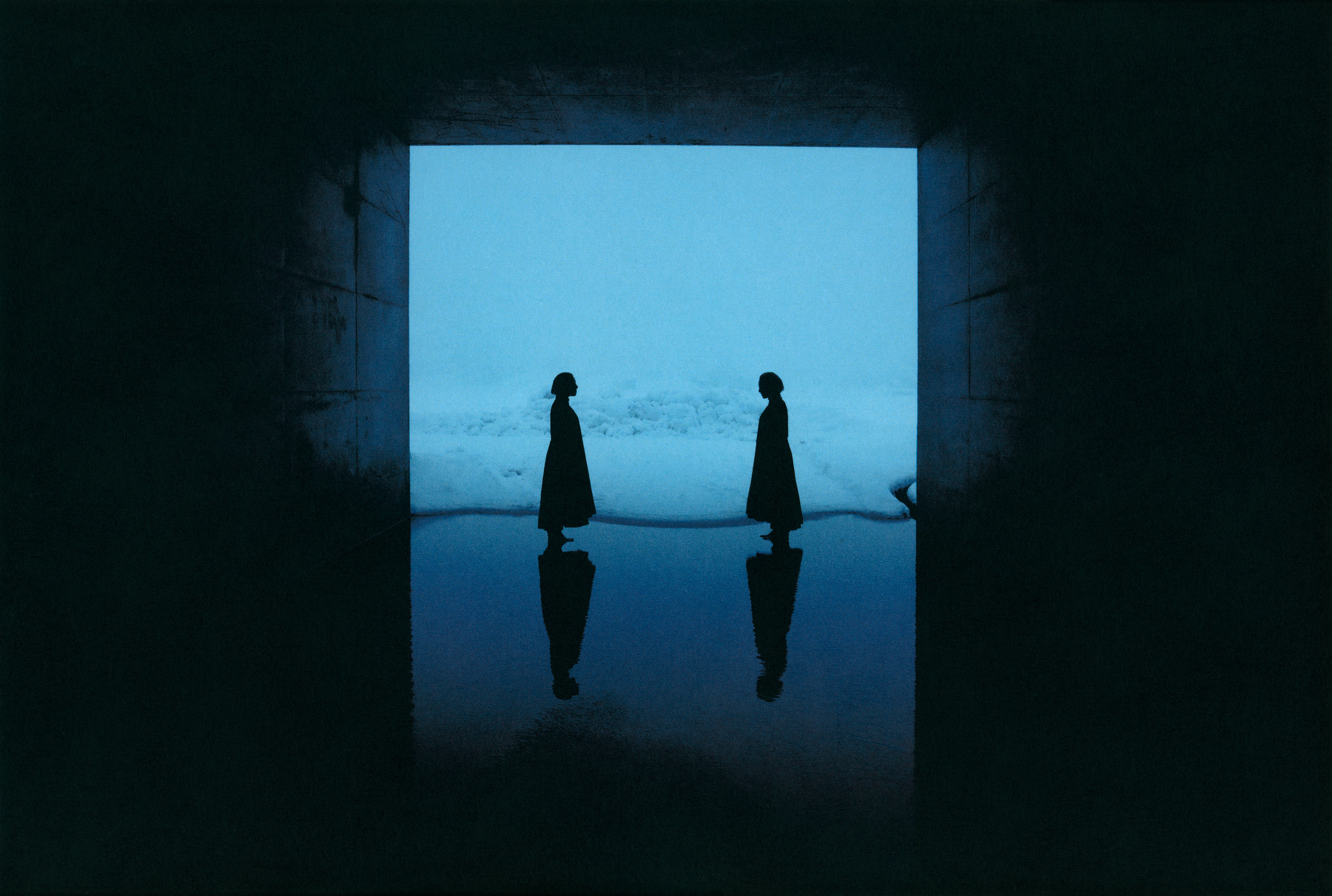 Colour photograph of two silhouetted figures standing face to face inside a dark tunnel, framed by a glowing blue expanse of snow and their own reflections on the wet ground.