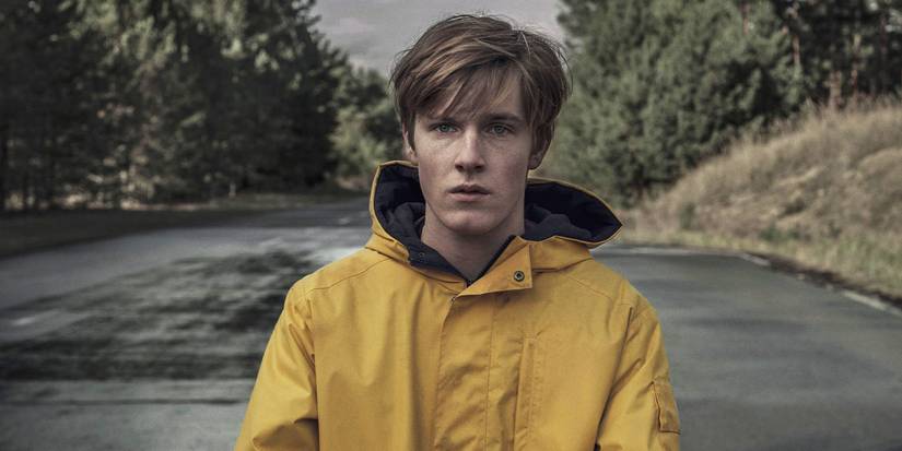 Louis Hofmann in a yellow raincoat standing on a deserted road in Dark.