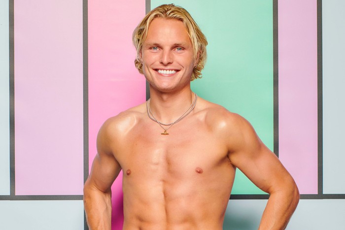 love-island (39) A topless man posing with his hands on his hips, smiling ahead in front of a colour block background.