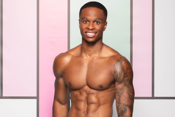 love-island (48) A topless man stood smiling ahead in front of a light green and pink panel background.
