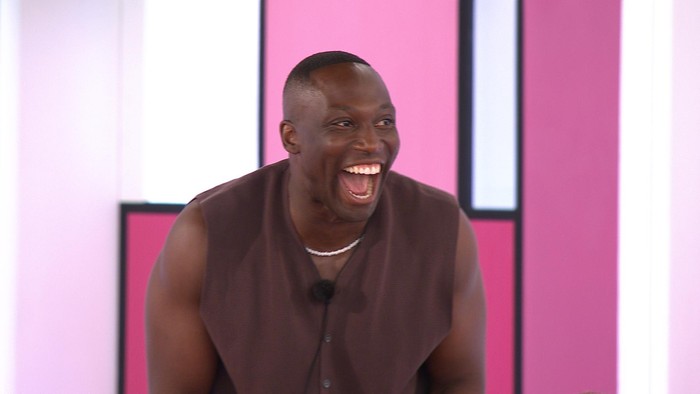 Love Island Ope slightly leant forward laughing and smiling while stood in front of a colour block background.