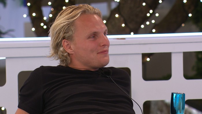 Love Island S13 A blonde man wearing a black t-shirt, smiling as he sits leant back.