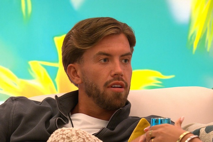 Love Island S13 Aidan sat back on a chair looking over at someone with a slight surprised expression.