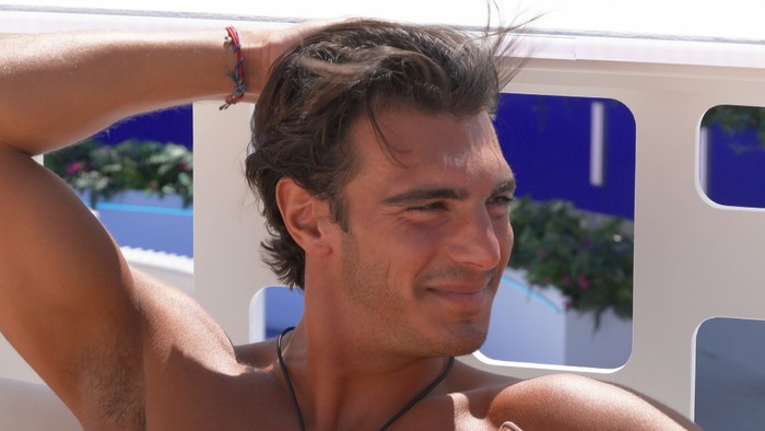 Love Island S13 Lorenzo sat on the terrace smiling as he looks over at someone.