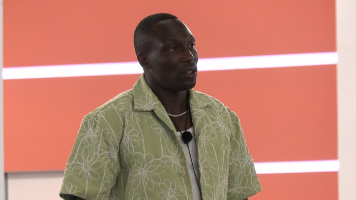Love Island S13 Ope wearing a white vest top and green shirt, looking ahead at someone.