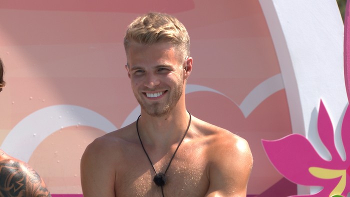 Love Island S13 Sam stood topless in the challenge arena, smiling as she looks over.