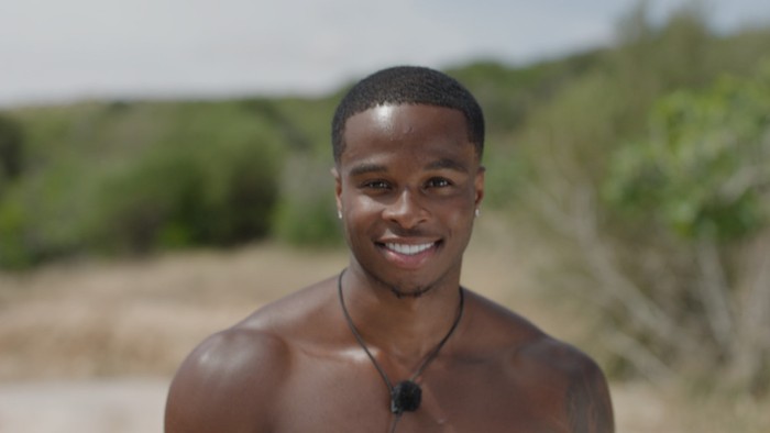 Love Island S13 A topless man smiling ahead in front of a green background.