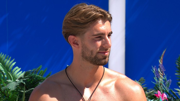 Love Island S13 A topless man looking over at someone while sat down, smiling.