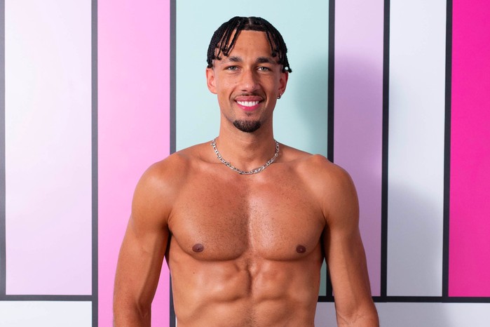 Love Island S13 Jordon stands in front of a colourful background and is topless. He has dark brown hair in plaits and is wearing a silver chain necklace.
