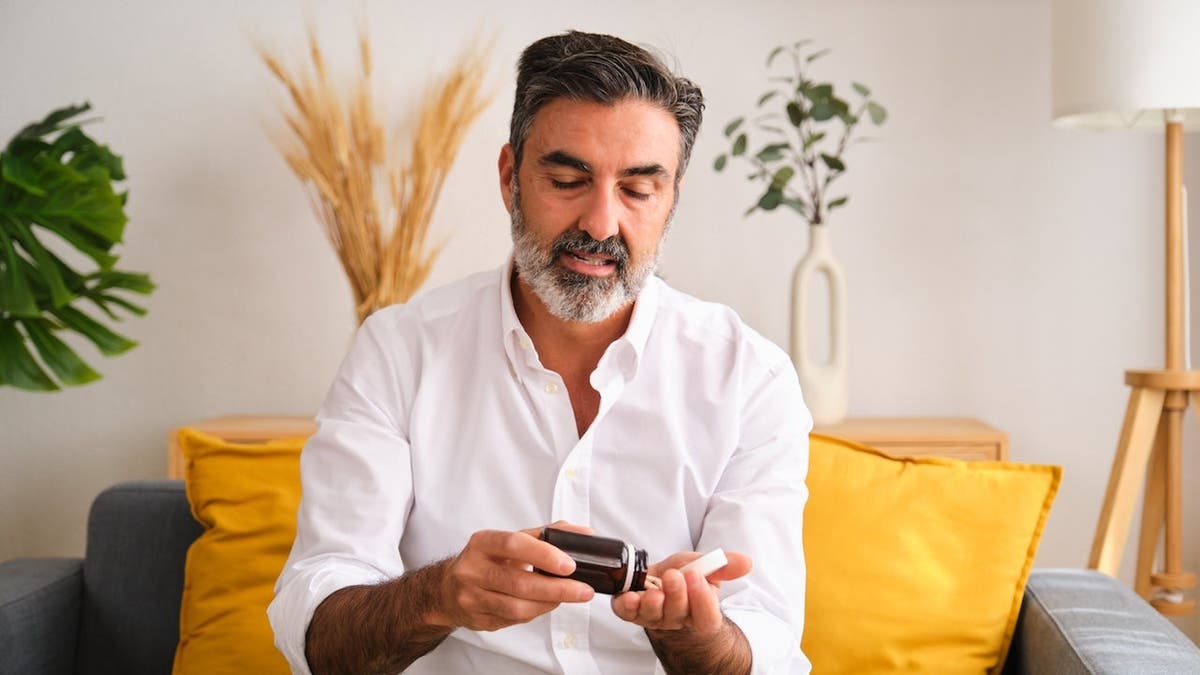 Older man in white shirt on couch taking supplements from bottle.