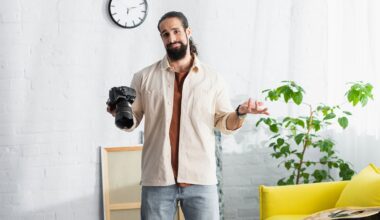 A man with a beard holds a camera in one hand and shrugs with the other, standing in a bright room with a yellow couch, green plant, and a clock on the wall.