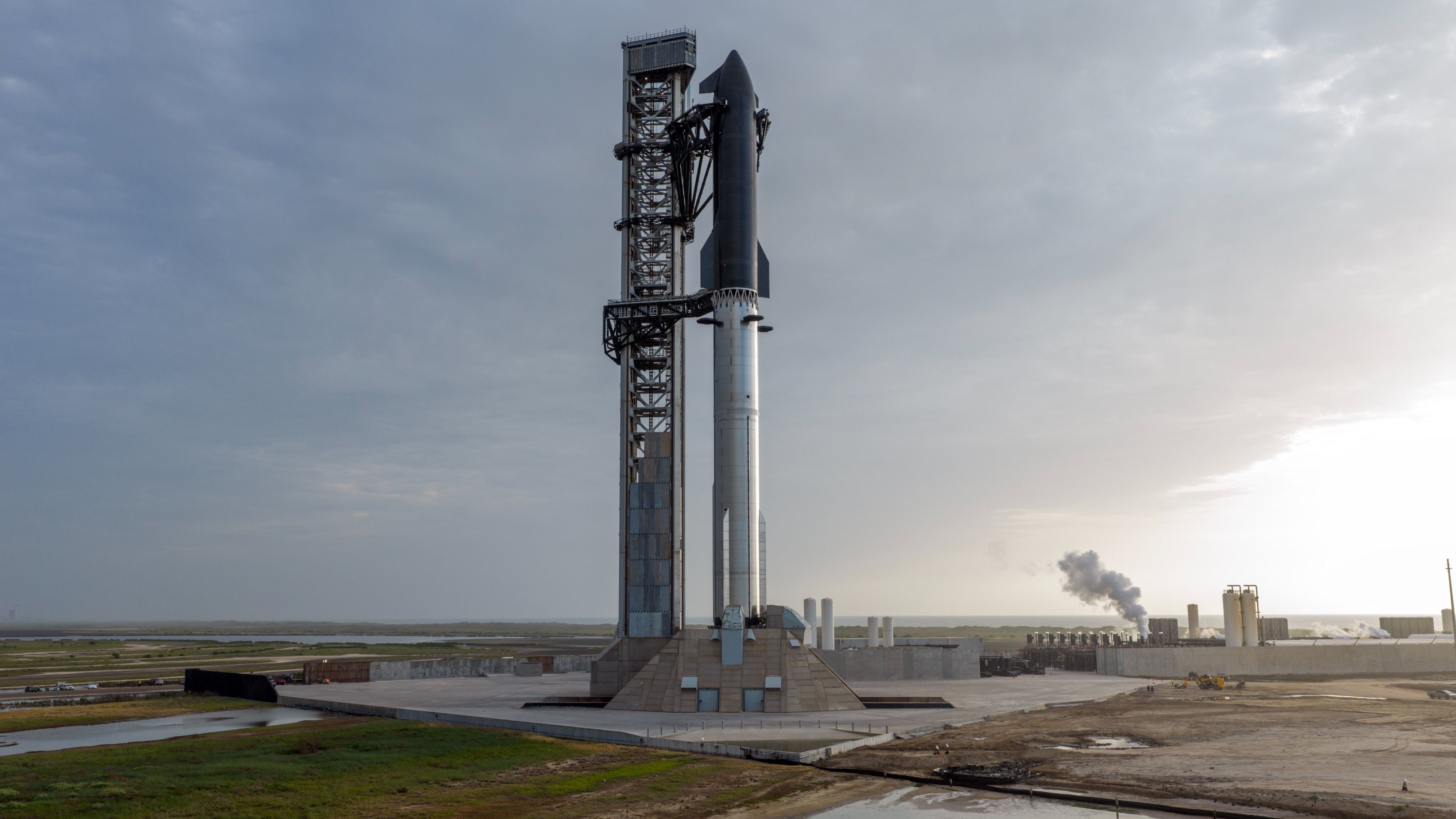 A giant steel rocket stands next to a launch tower against a cloudy sky