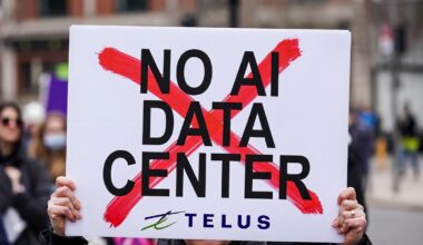 Protest sign reading 'NO AI DATA CENTER' with a red X behind the text and a Telus logo at the bottom, held by hands in a crowd.