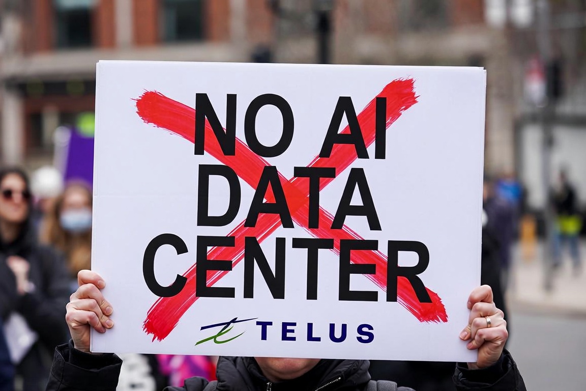 Protest sign reading 'NO AI DATA CENTER' with a red X behind the text and a Telus logo at the bottom, held by hands in a crowd.