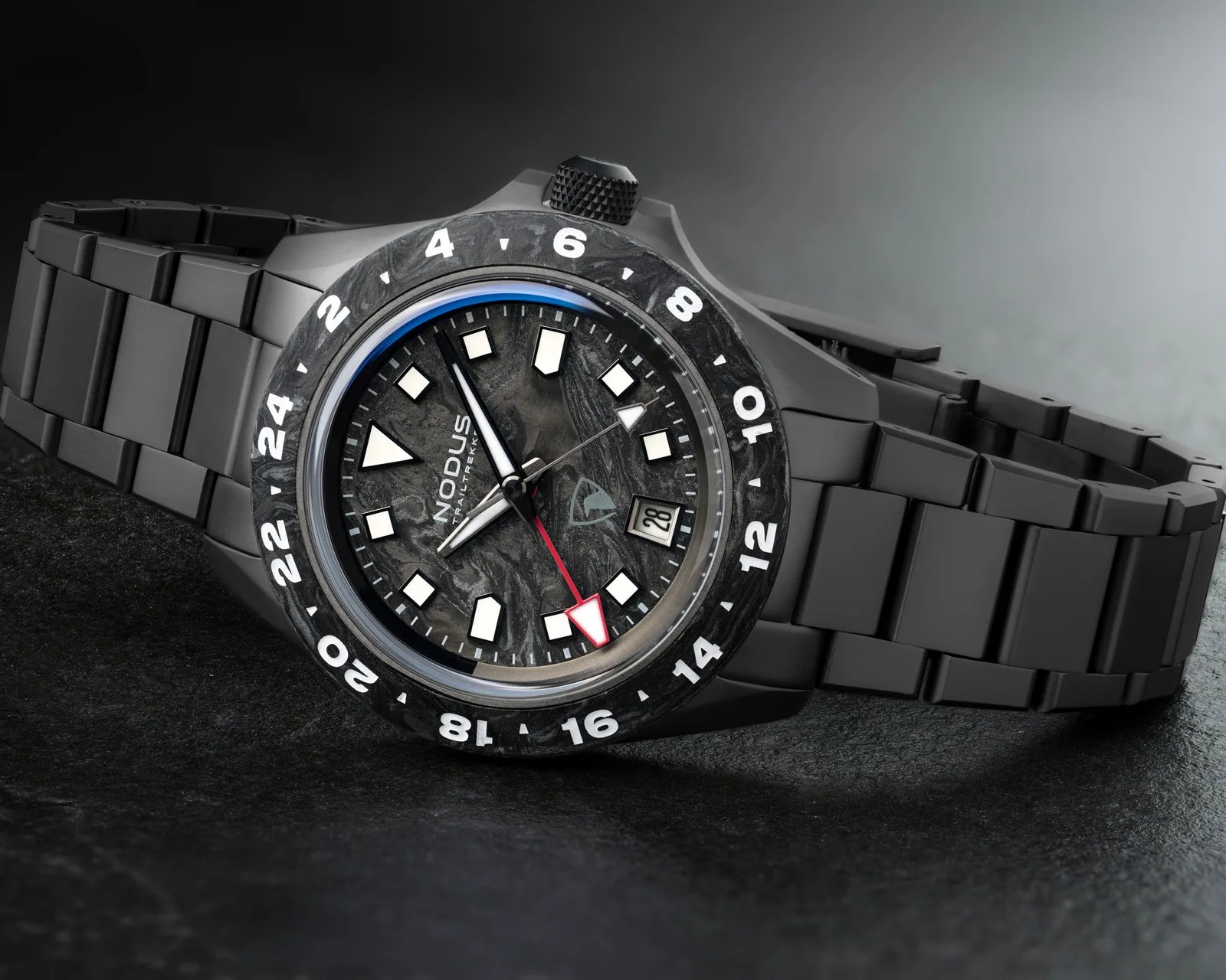 Black metal wristwatch with a textured dark dial, white hour markers, red-tipped GMT hand, and date window on a black surface.