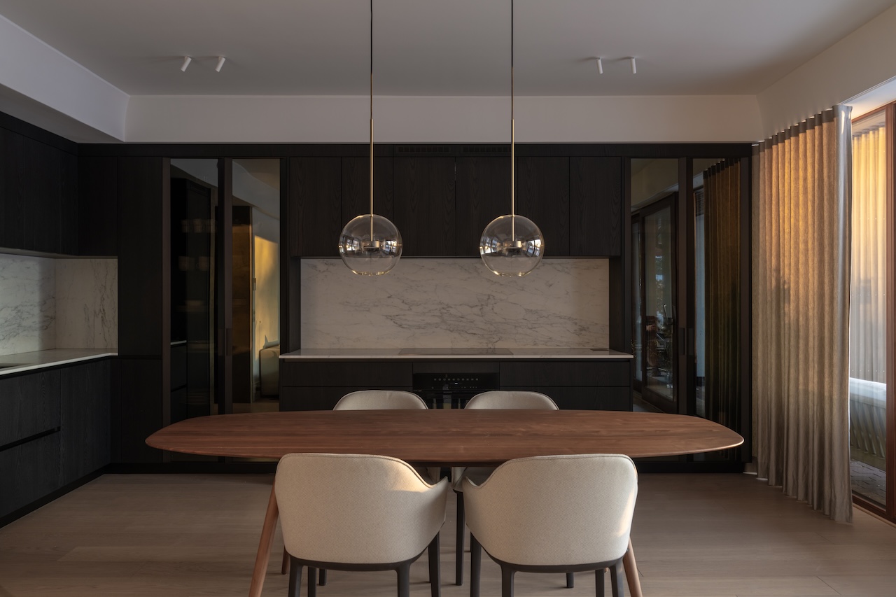 Modern dining area with a wooden table, four upholstered chairs, dark cabinetry, marble backsplash, and two spherical pendant lights. Floor-to-ceiling window with sheer curtains on the right.
