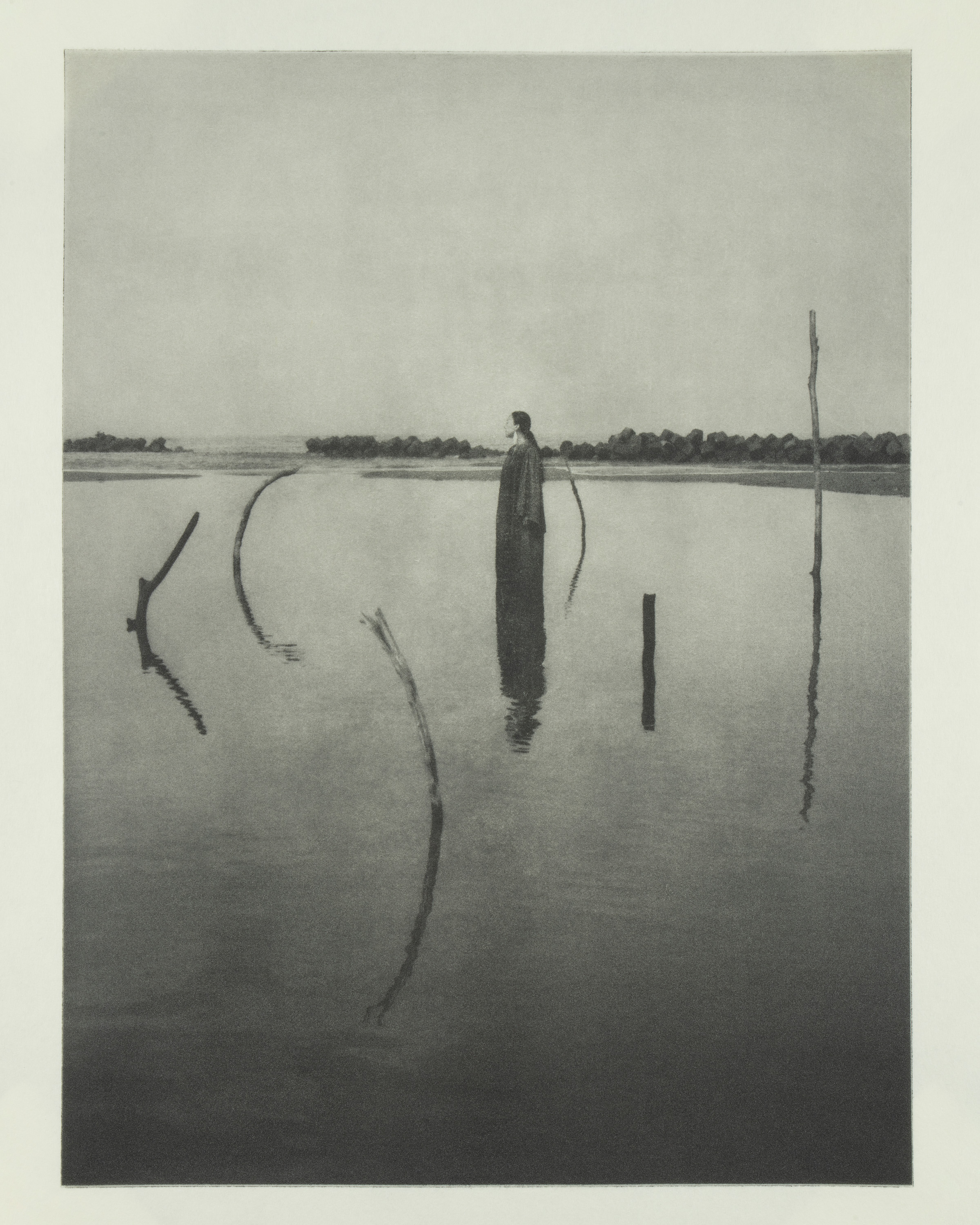 Black and white photograph of a solitary figure standing in a wide, calm body of water, surrounded by thin curved sticks that break the surface.