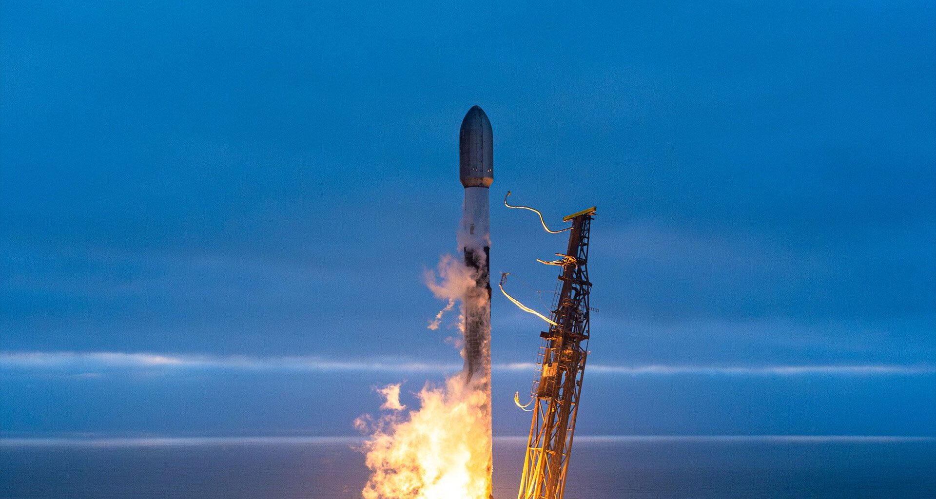 A white and black rocket launches into an overcast evening sky from an ocean-side pad.