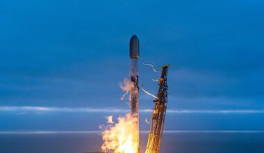 A white and black rocket launches into an overcast evening sky from an ocean-side pad.