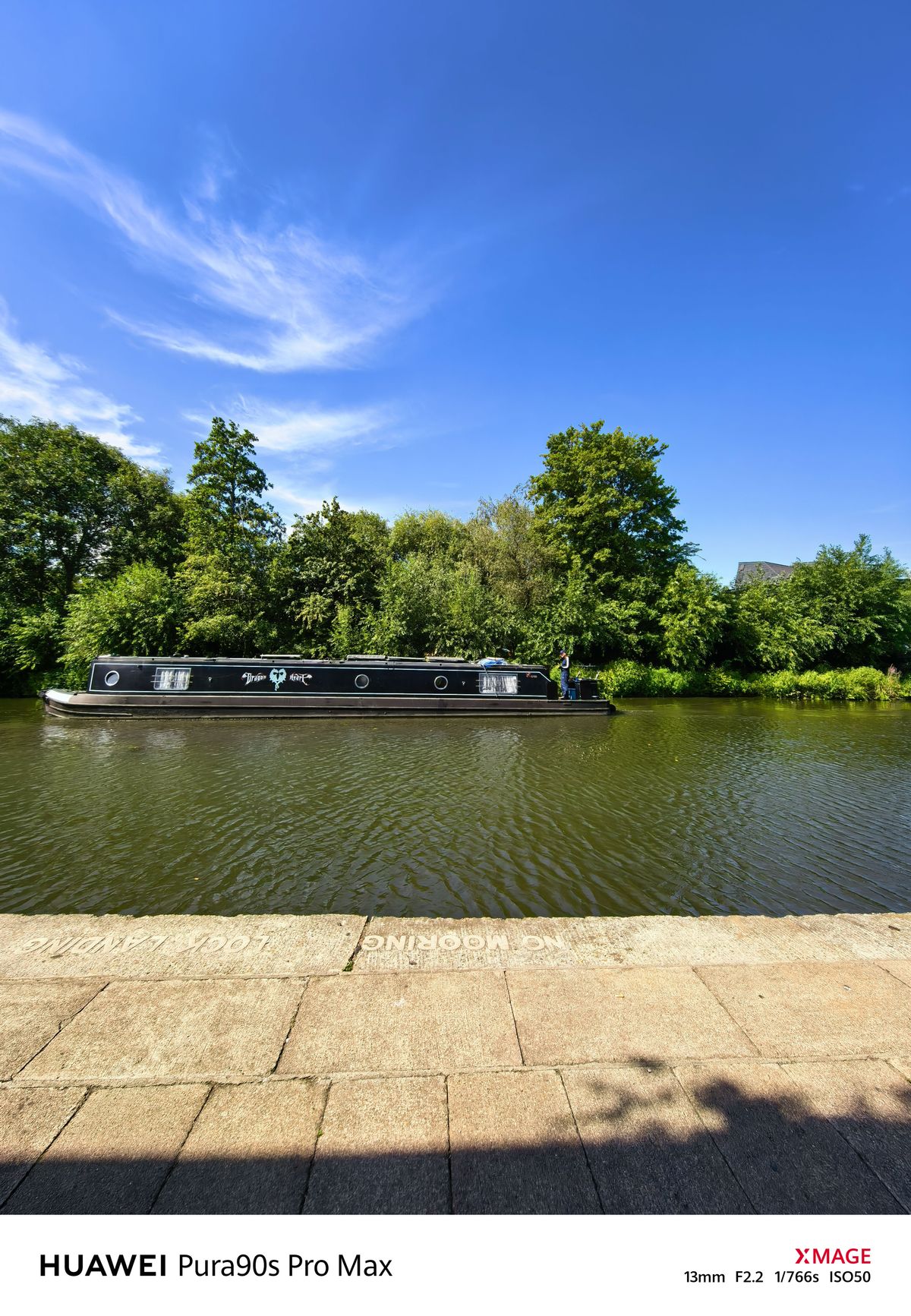 Long black narrowboat traveling along a canal bordered by dense green trees under a bright blue sky.