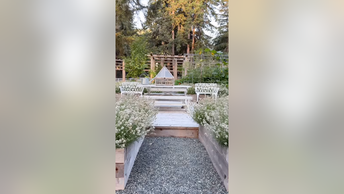 White benches and a picnic table sit beside raised flower beds and a small greenhouse.