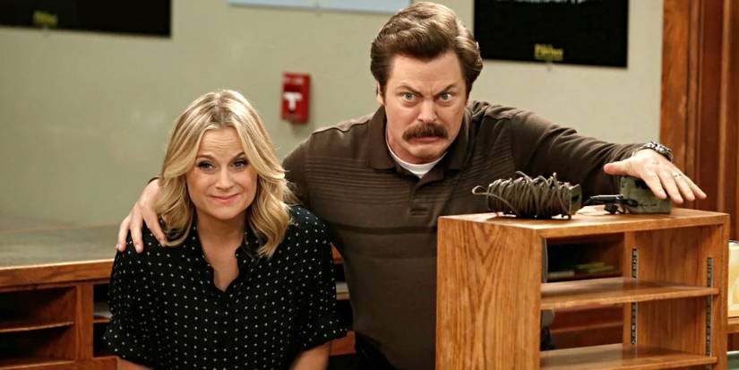 Amy Poehler and Nick Offerman stand behind a bookshelf and look concerned in Parks and Recreation.