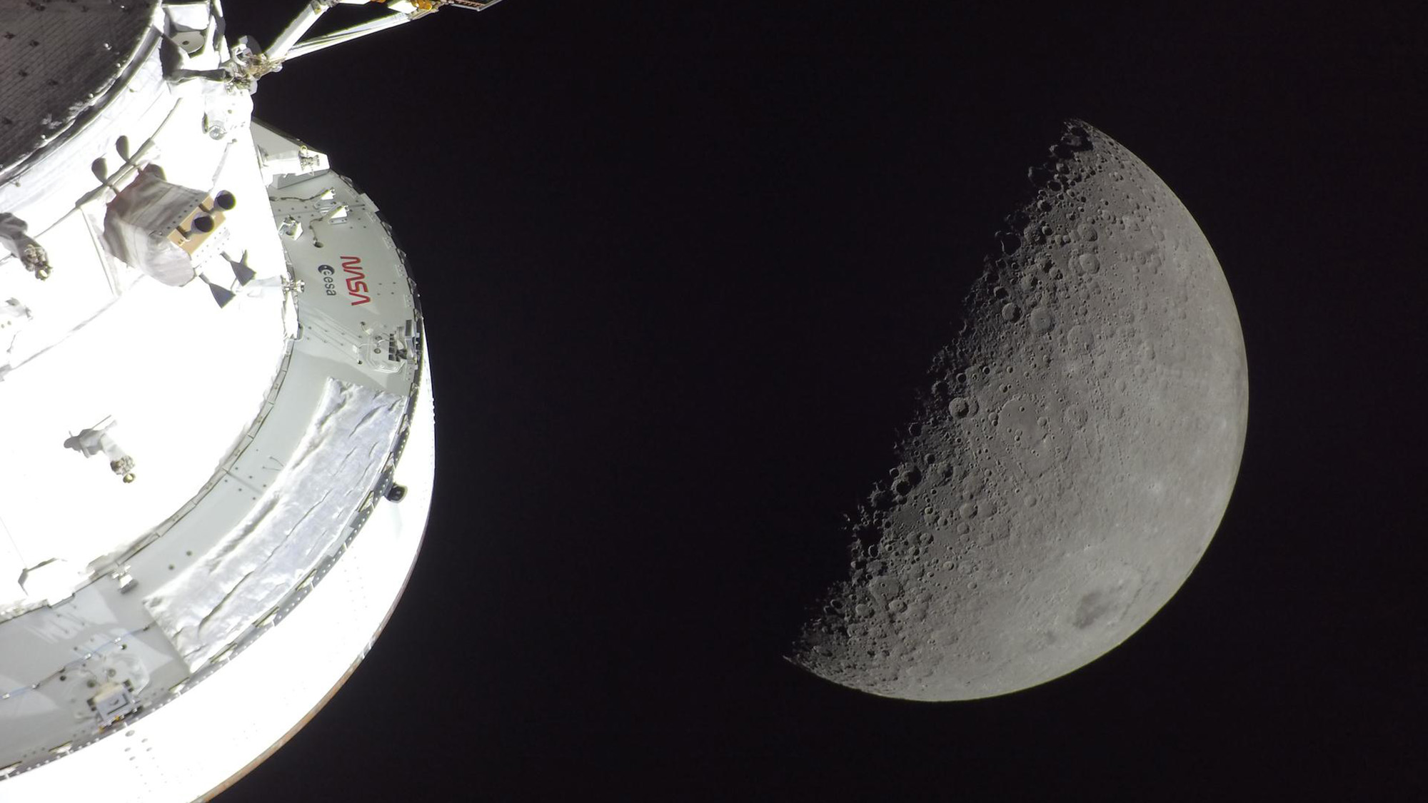A view of the gray moon with a white spacecraft in the bottom left of the image.