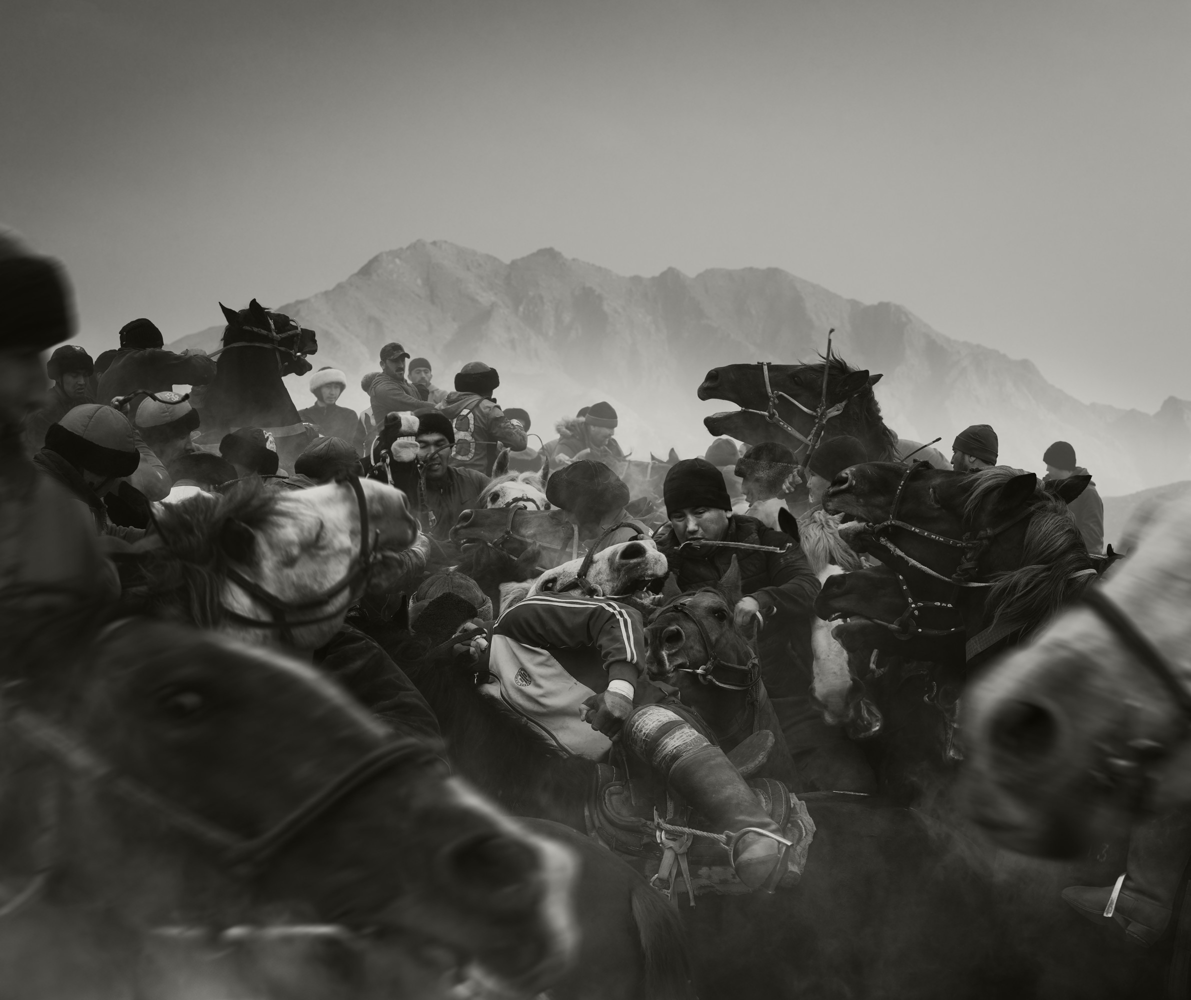 A dramatic, black-and-white action photograph captures a dense crowd of men on horseback closely competing in a traditional, dusty game of Buzkashi with mountains visible in the hazy background