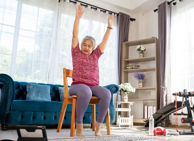 Senior woman sitting on chair and raising two arms above her head. doing workout for good healthy with training exercise online on tablet, elderly workout online from home concept