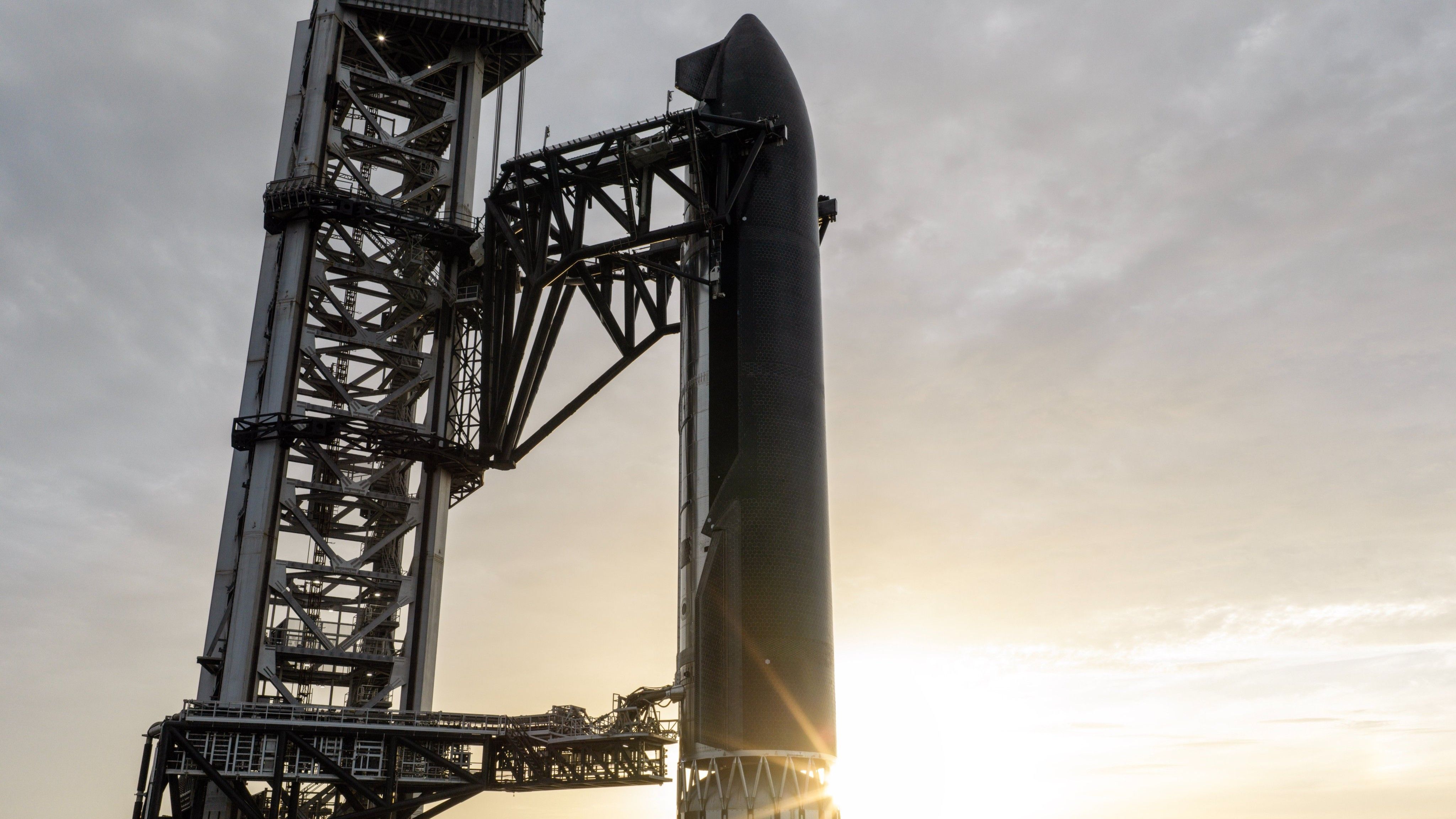 A giant steel rocket stands next to a launch tower with the sun beaming from below