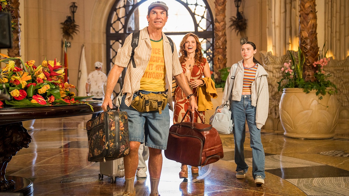 Jerry O'Connell walks through a hotel lobby carrying luggage with his co-stars, Violet McGraw and Sophia Bush.