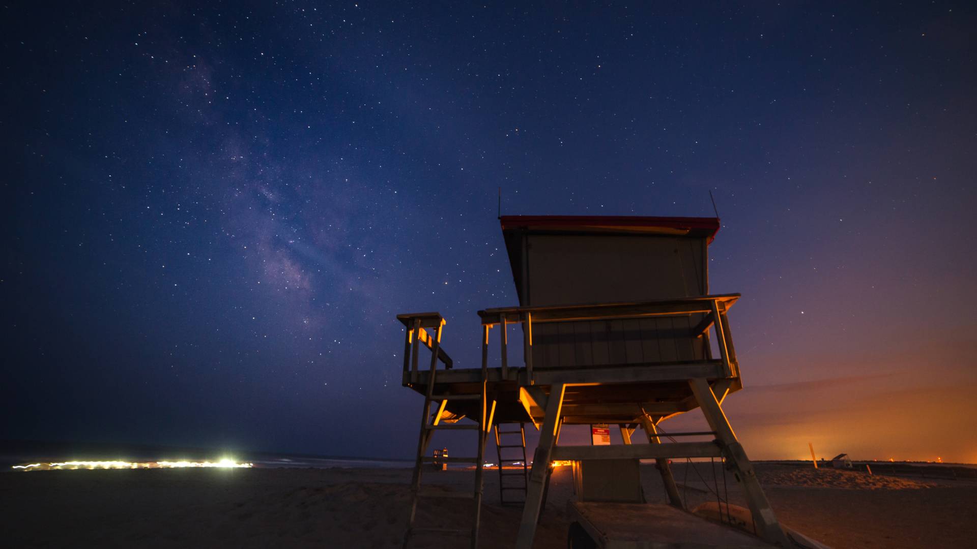 A night sky filled with stars and the Milky Way. In the foreground is a beach and a wooden lifeguard stand.