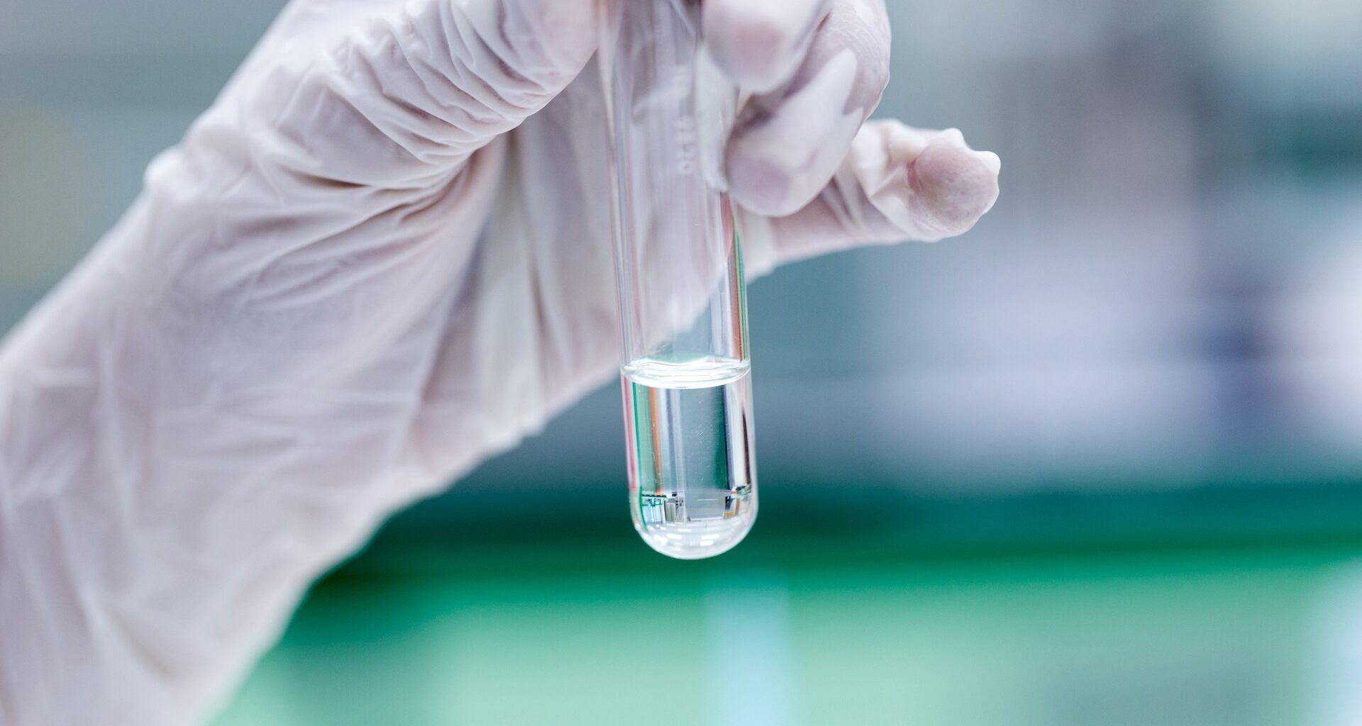 A gloved hand holds a test tube containing a clear liquid against a blurred laboratory background.