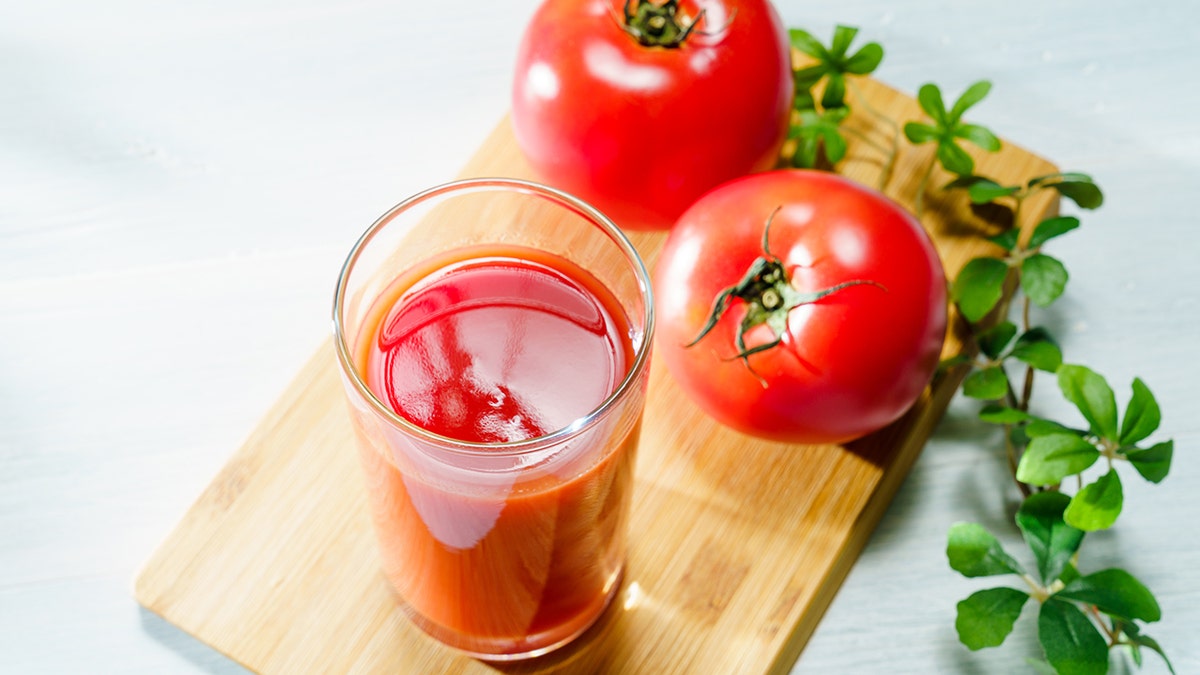 A glass of tomato juice and two tomatoes on a wooden cutting board.