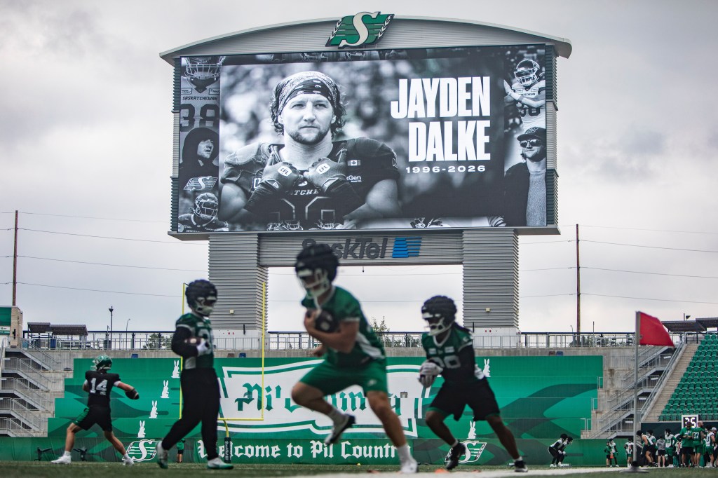 Football players practice on a field under a scoreboard displaying a tribute to late Roughriders player Jayden Dalke.