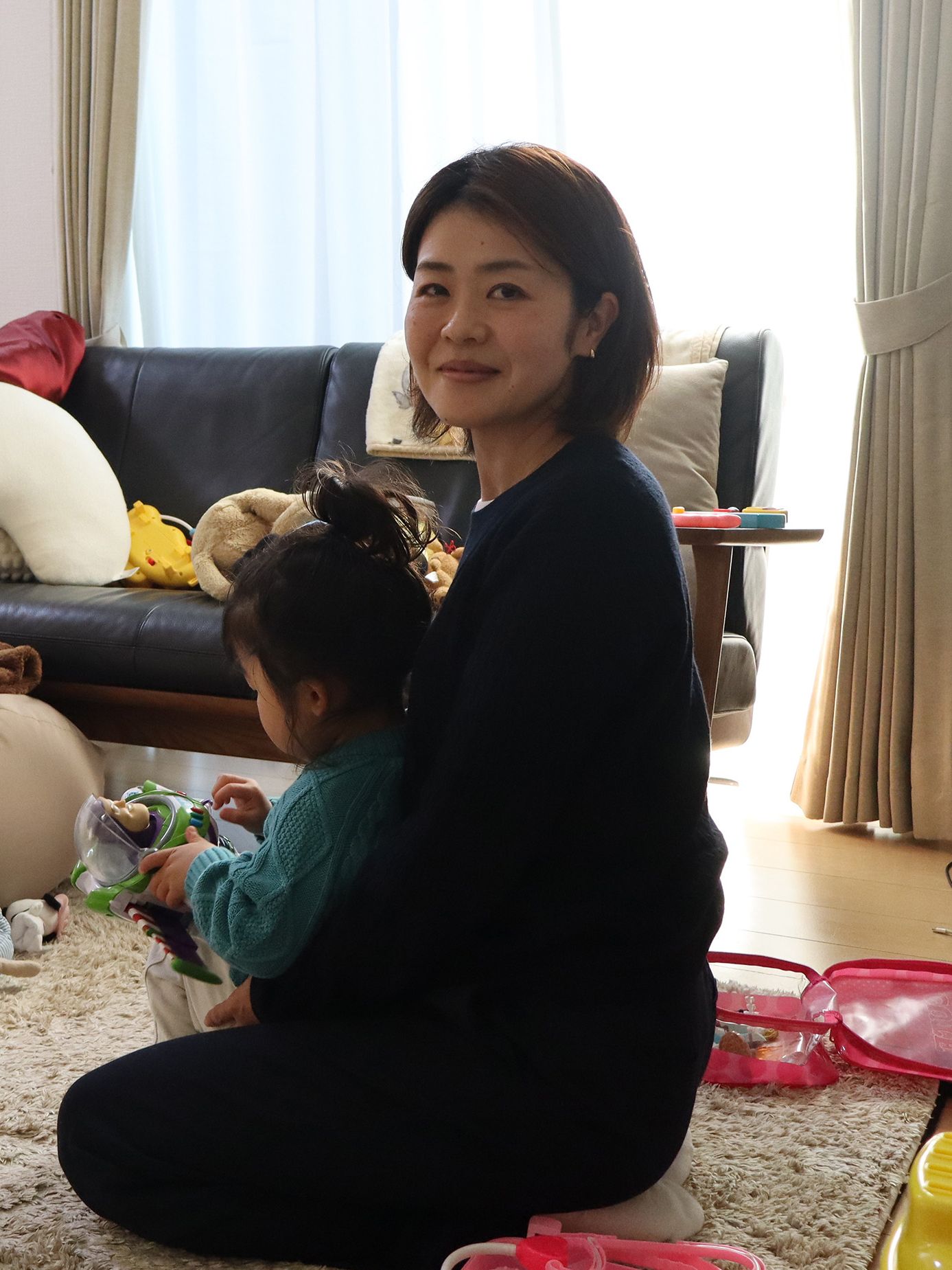a photo of a smiling woman with short hair sitting on the floor of her home, holding a toddler who in turn is playing with a Buzz Lightyear action figure