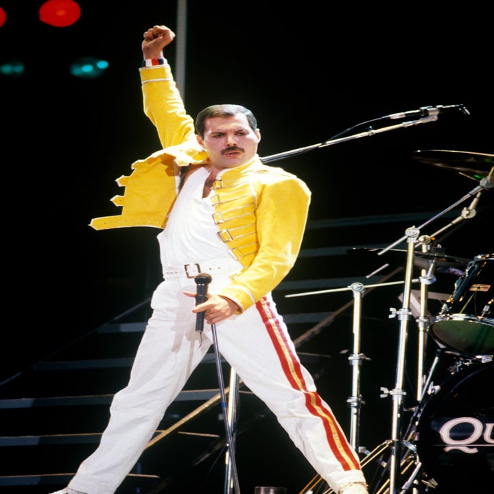 Musician on stage in a yellow jacket, white pants with red stripes, striking a dynamic pose with a microphone stand