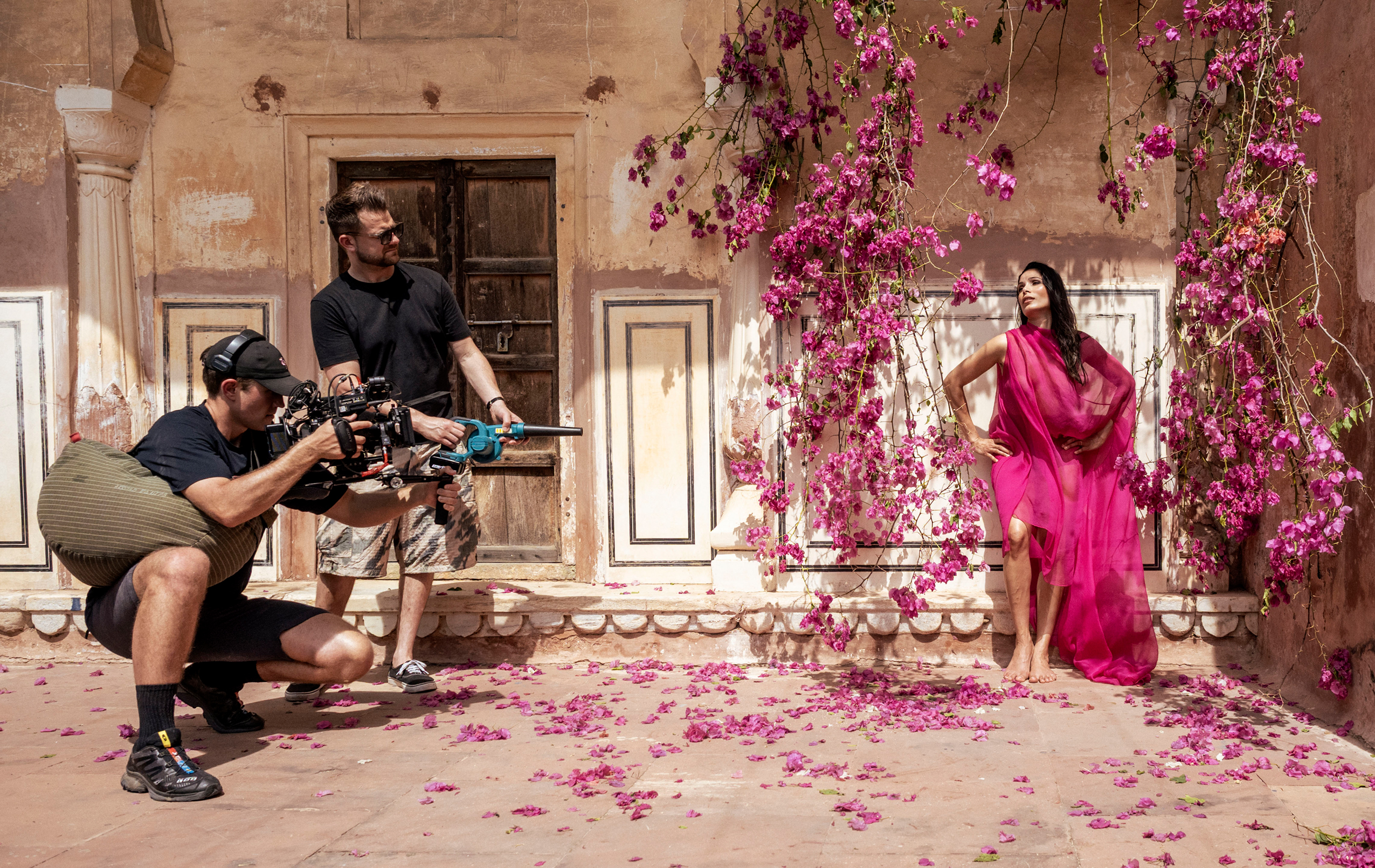 A cream colored backyard with the only color visible pink as pink flowers appear to the right where a woman poses in a pink dress, right is the video and photo crew capturing her