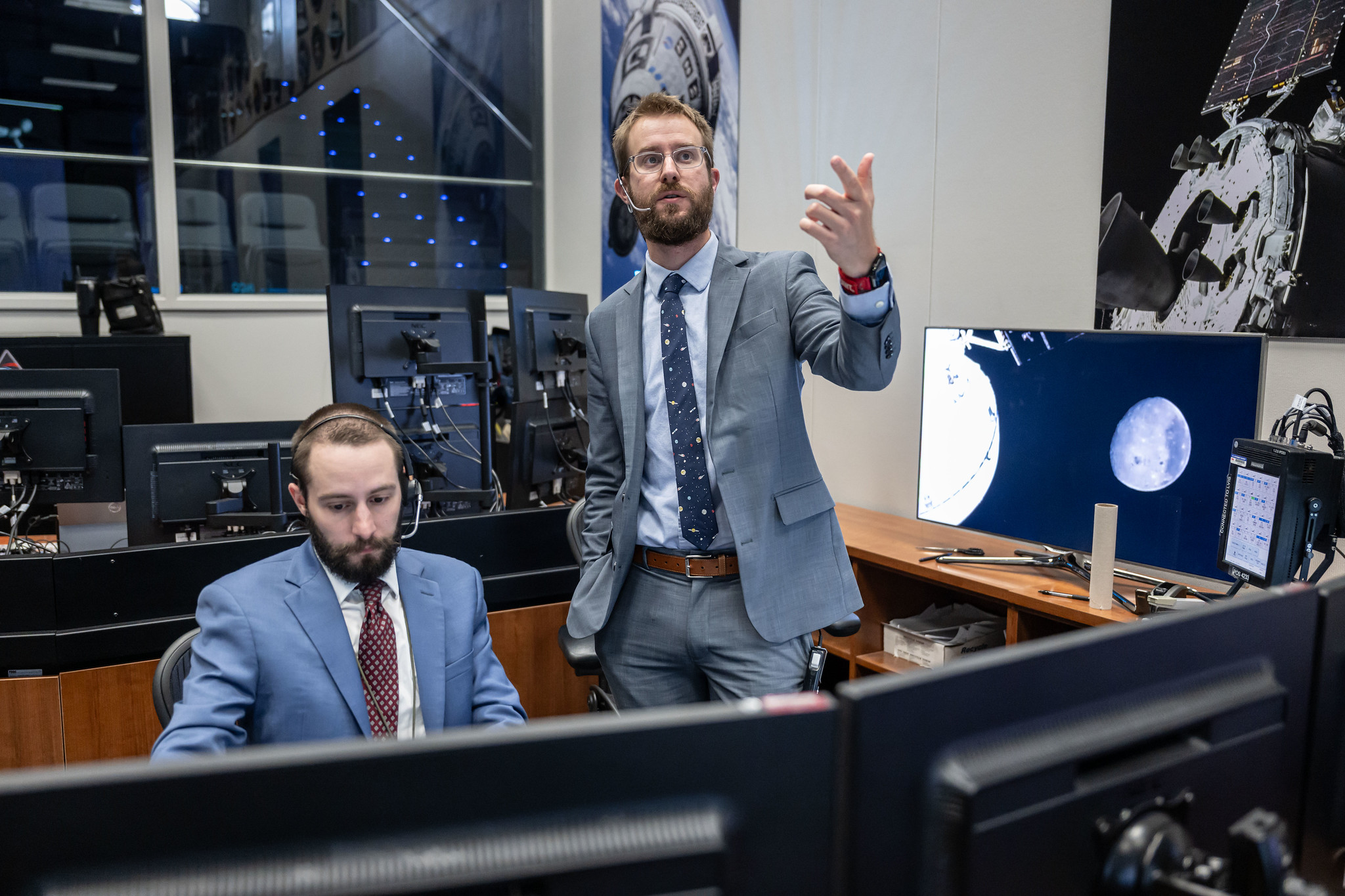 Two men in blue and gray suits and ties wearing headsets look at several monitors.