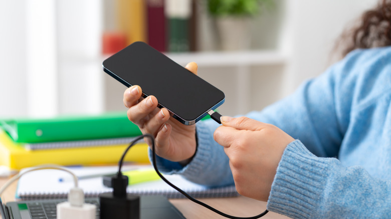 A person plugging a phone into a charger at their desk.