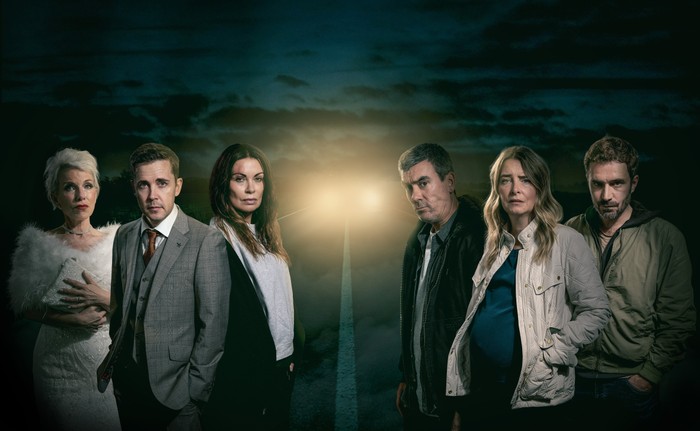 An image depicting a nighttime scene on a road with car headlights in the distance, from left to right we see Sue Devaney as Debbie Webster in a wedding dress, Gareth Pierce in a suit as Todd Grimshaw, Alison King as Carla Connor with a bloodied forehead, Jeff Hordley as Cain Dingle, Emma Atkins as a pregnant Charity Dingle, and Oliver Farnworth as a brooding John Sugden, in a promo image for Corriedale.
