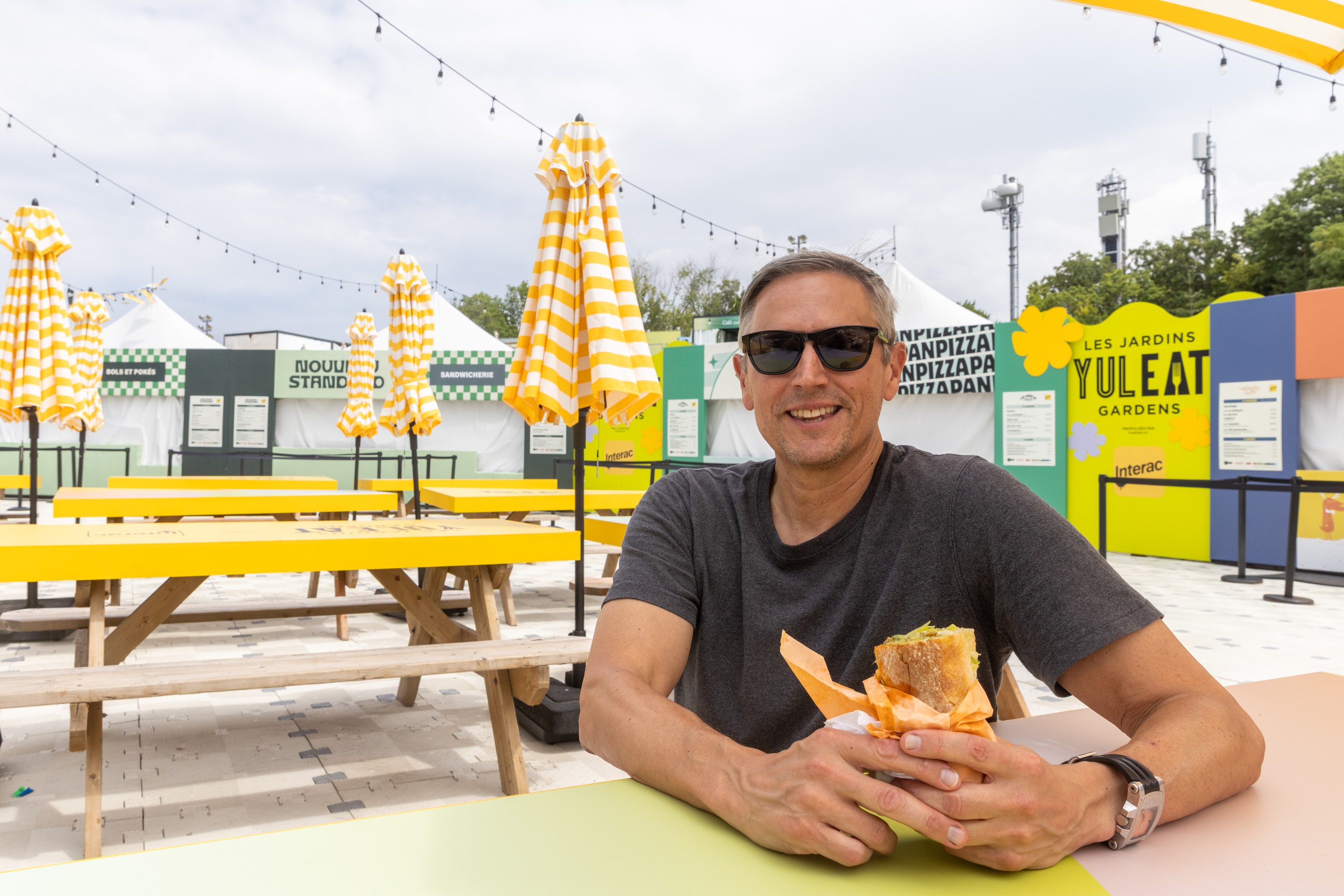 Man sits at table on festival site with submarine sandwich in his hands.