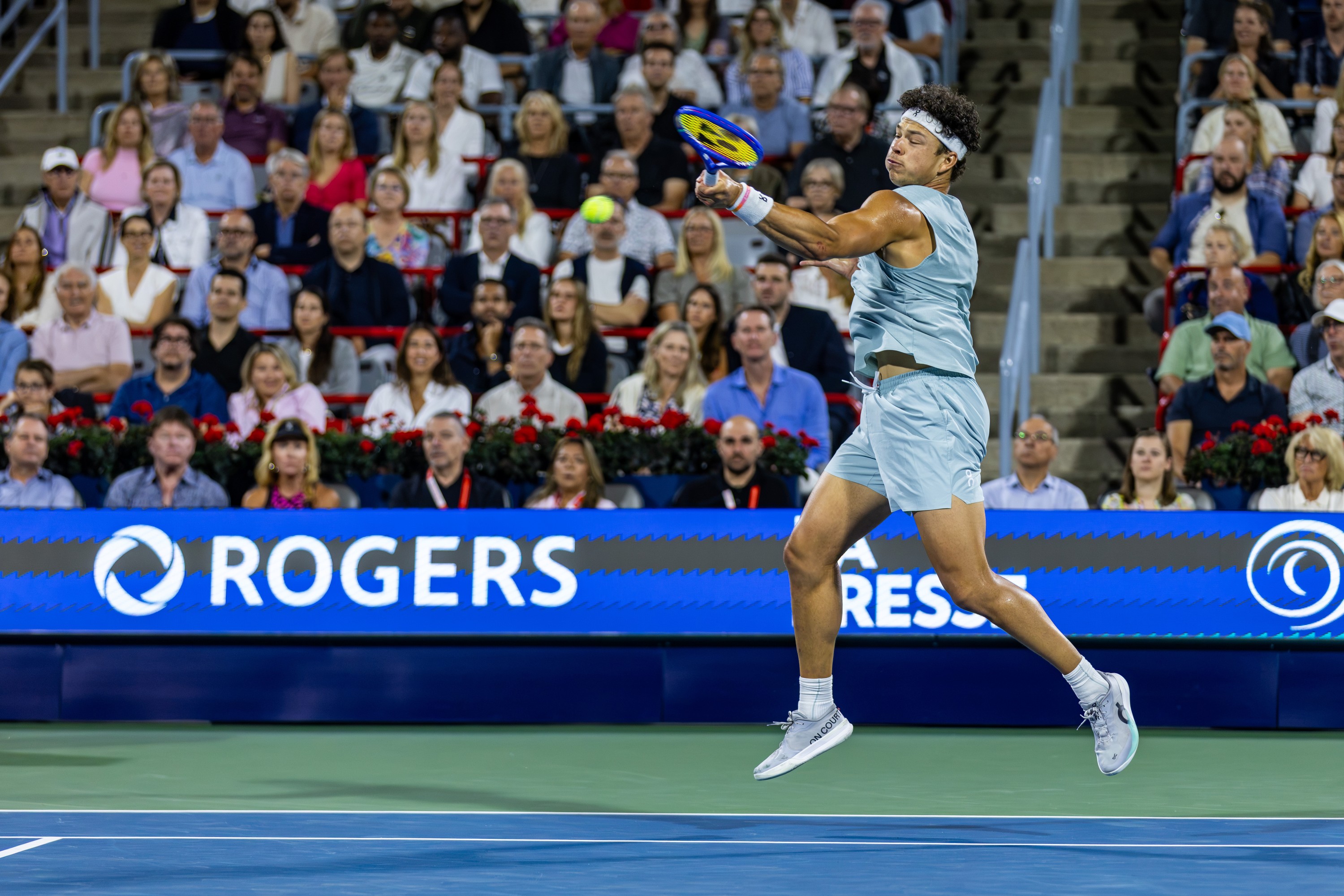 Ben Shelton's feet are off the ground and his arm outstretched to hit a tennis ball. Spectators are in the background.