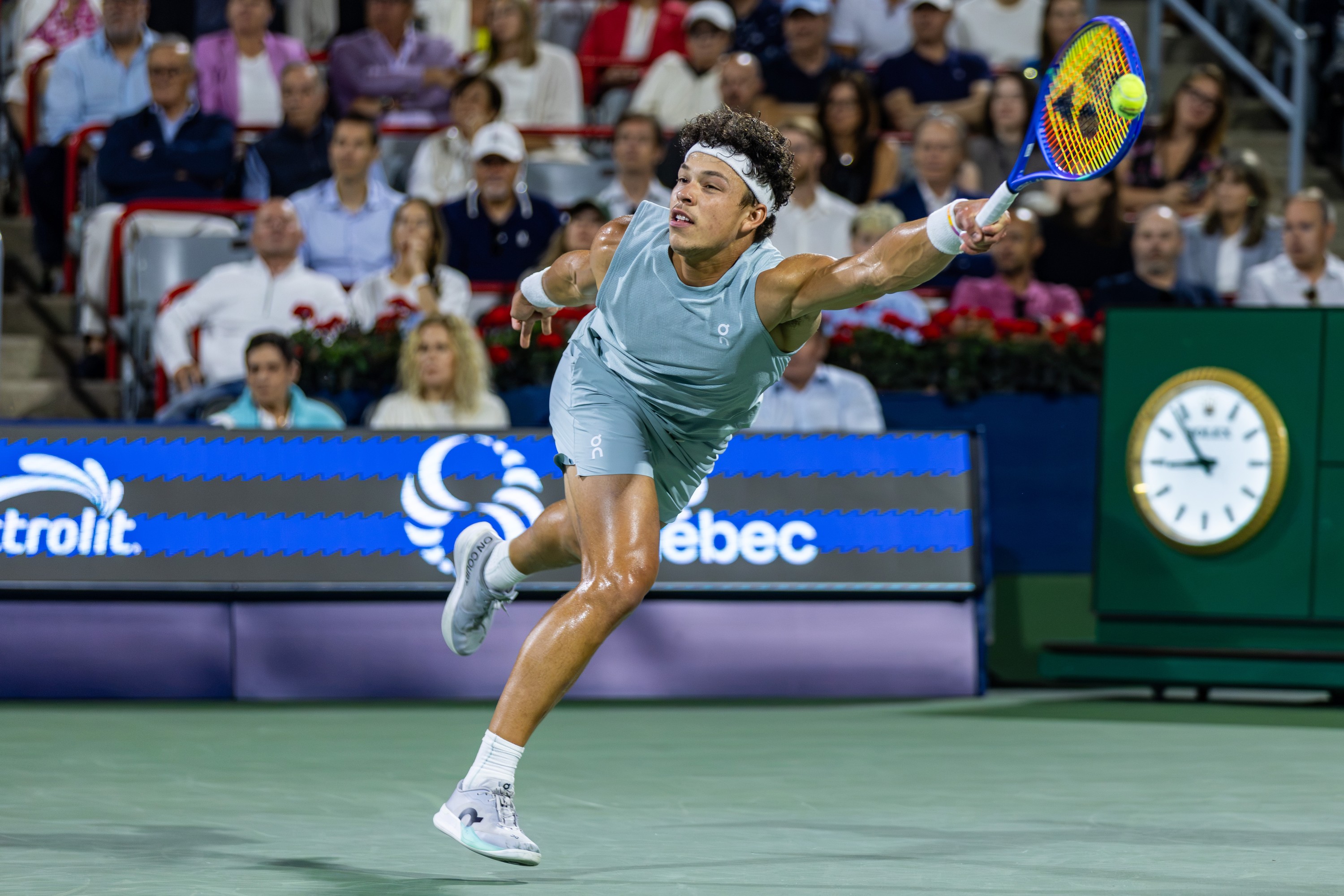 Ben Shelton leans far forward, racket arm outstretched, to hit a tennis ball.