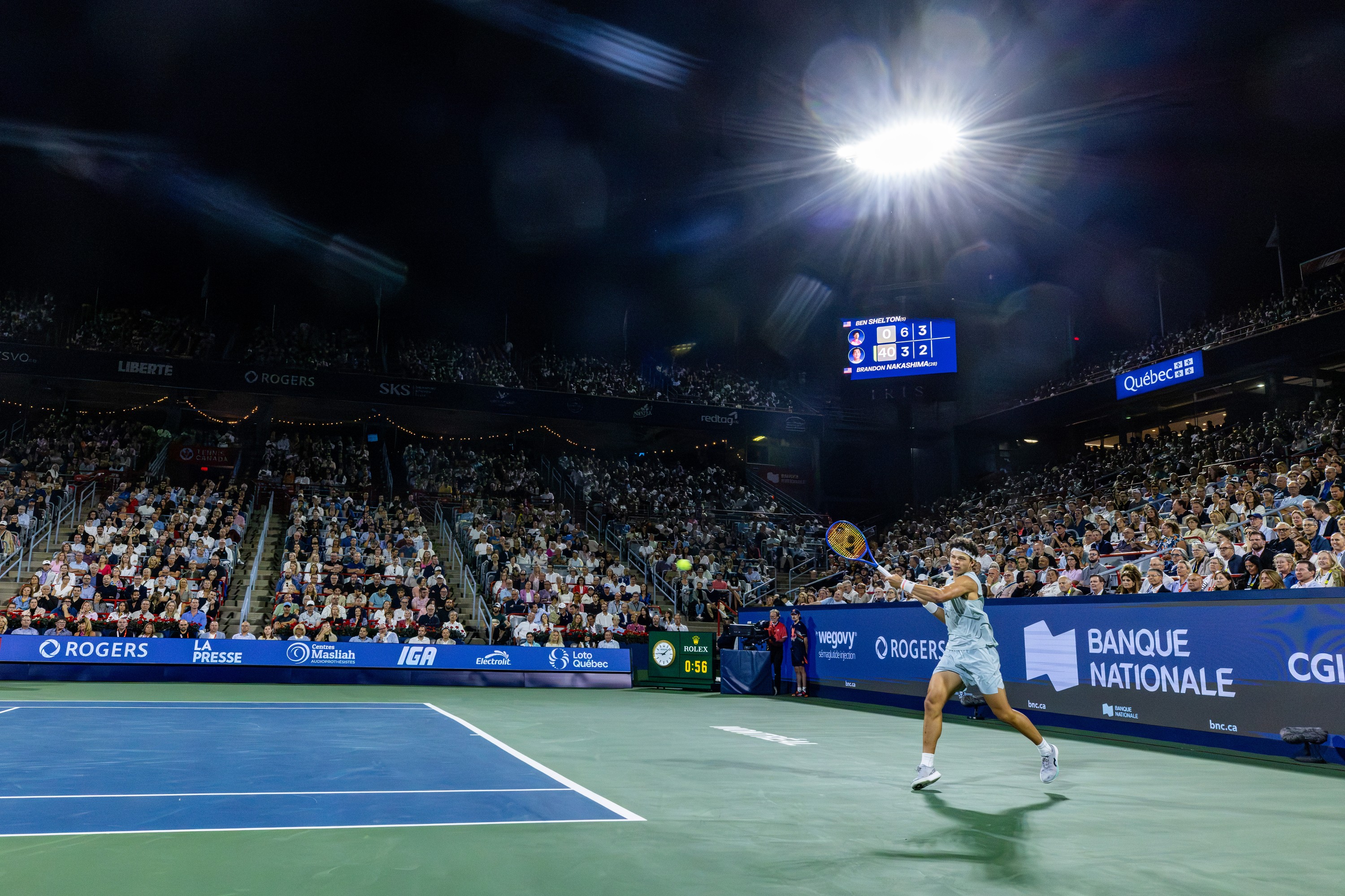A spotlight is shining on Ben Shelton as he hits a forehand shot, with audience members in the background.
