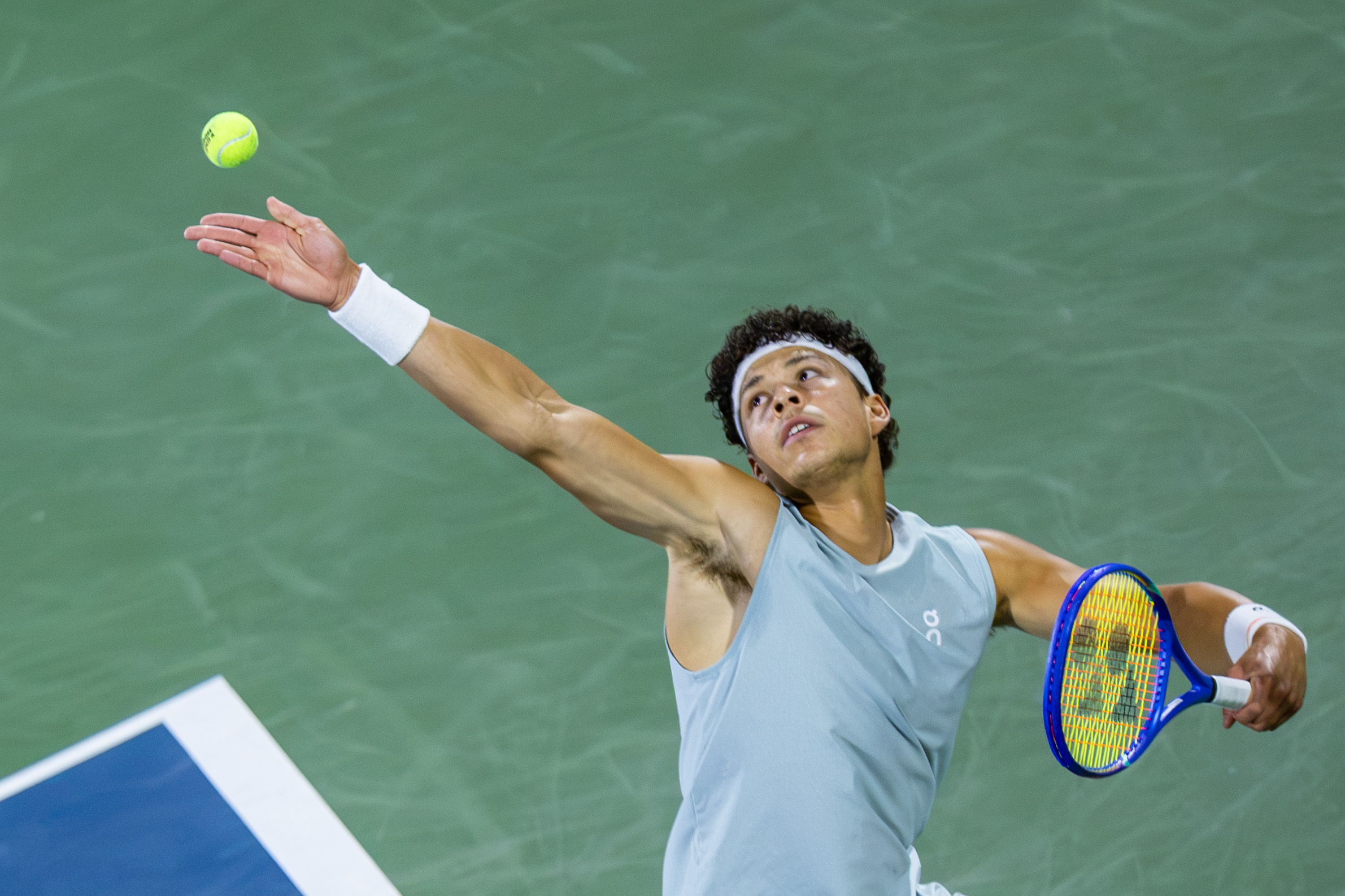 Ben Shelton's arm is stretched up and out as he serves a tennis ball.