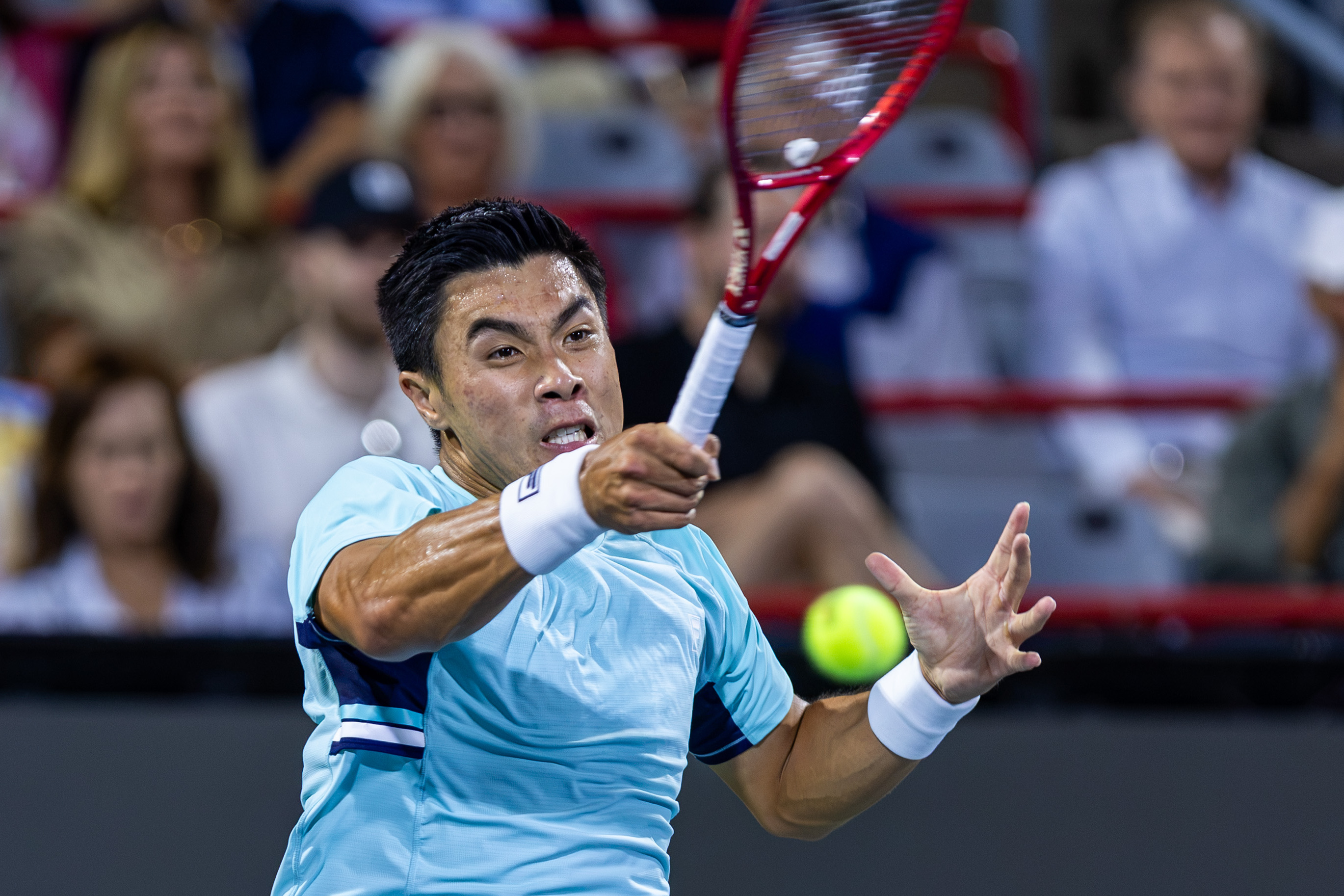 Brandon Nakashima hits a tennis ball, with spectators blurred behind him.