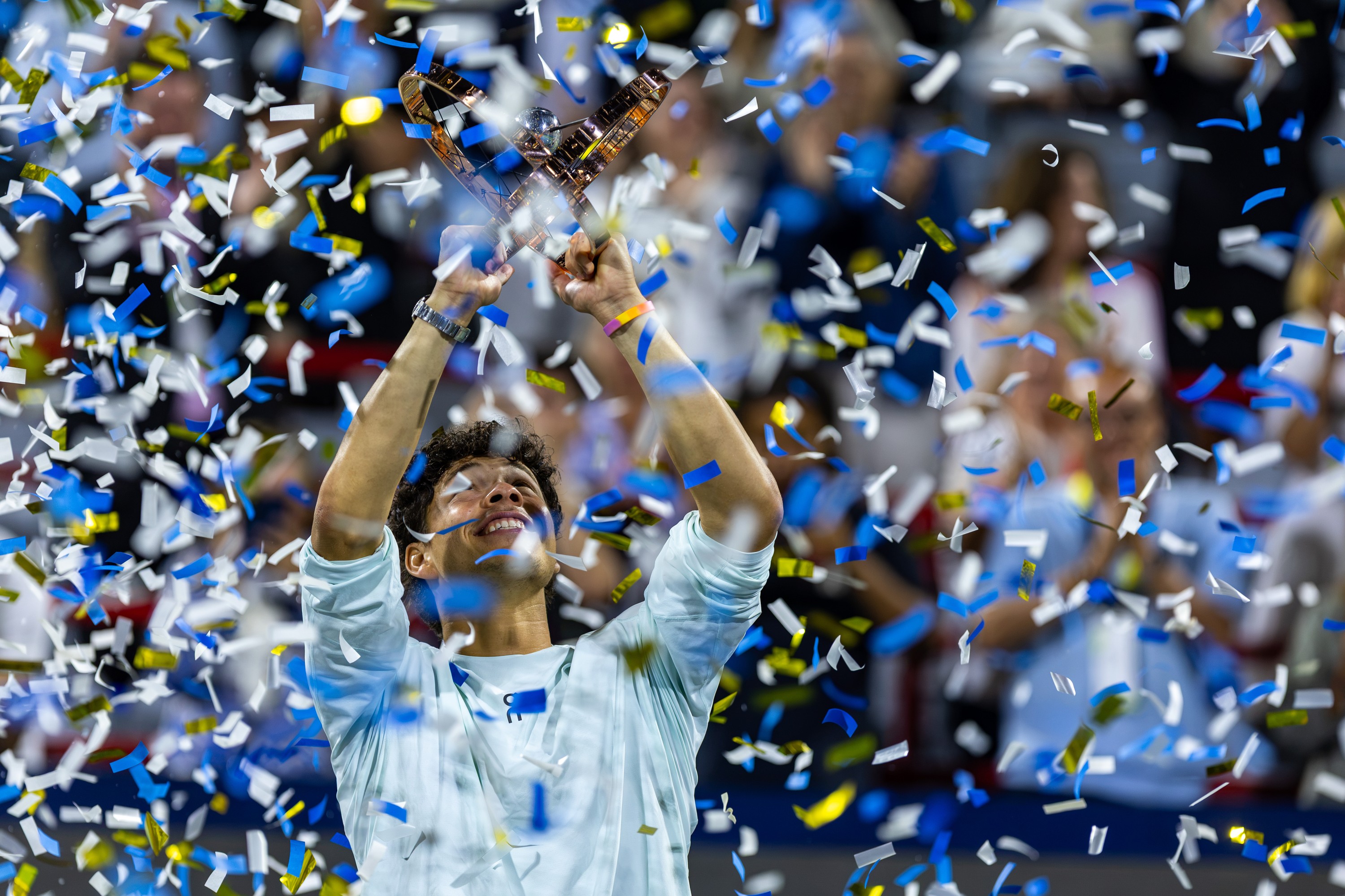 Ben Shelton, obscured by confetti, smiles widely and holds his trophy over his head.