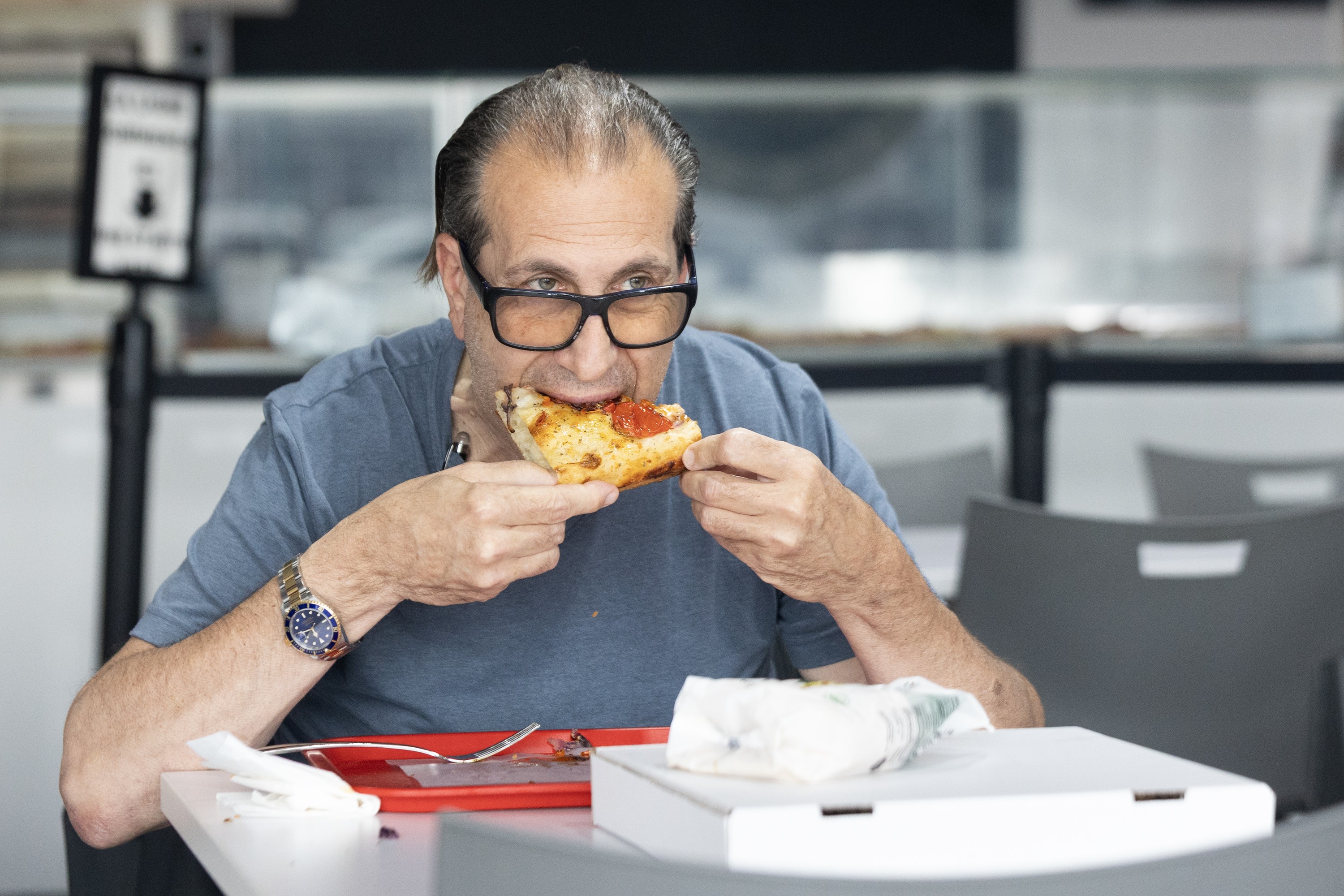Customer bites into a slice of al taglio pizza at Redipizza. A white pizza box is on the table.