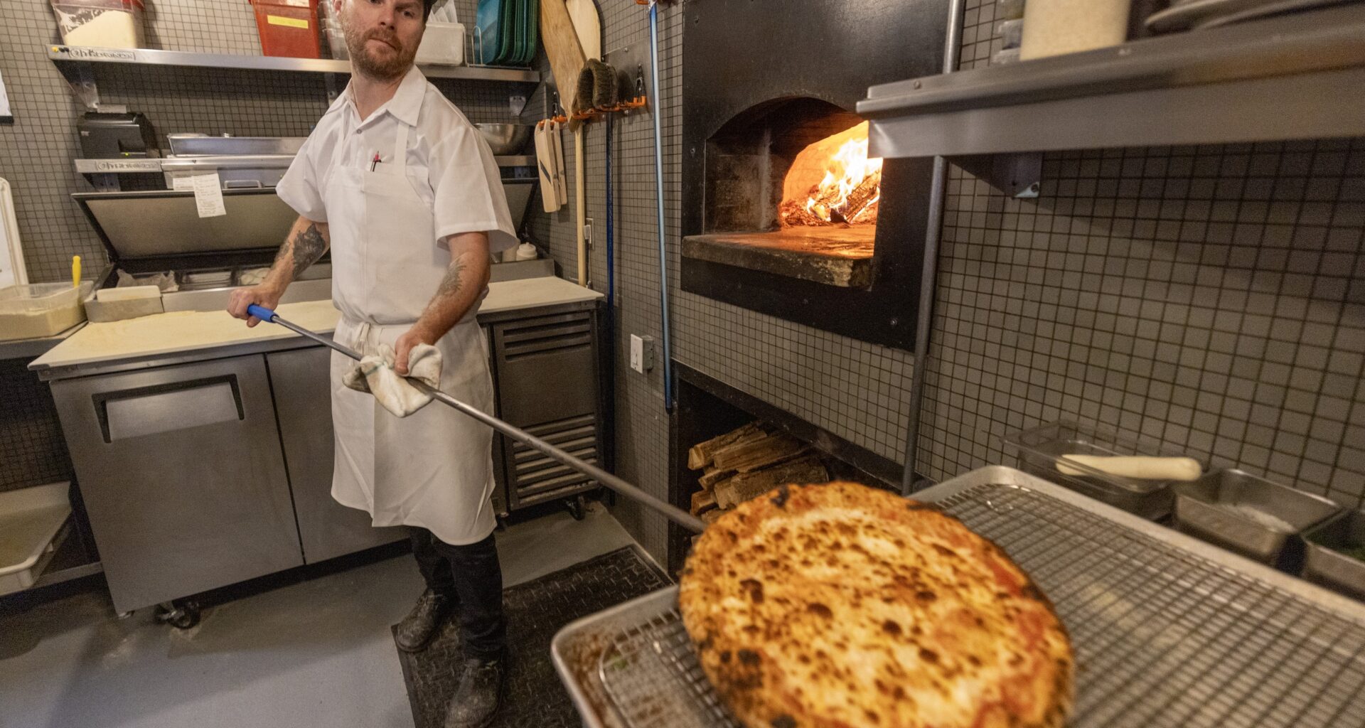 Restaurant executive chef Christopher Cameron, holds a pizza paddle with a cooked pizza on it, with a glowing wood-fired pizza oven to his right.
